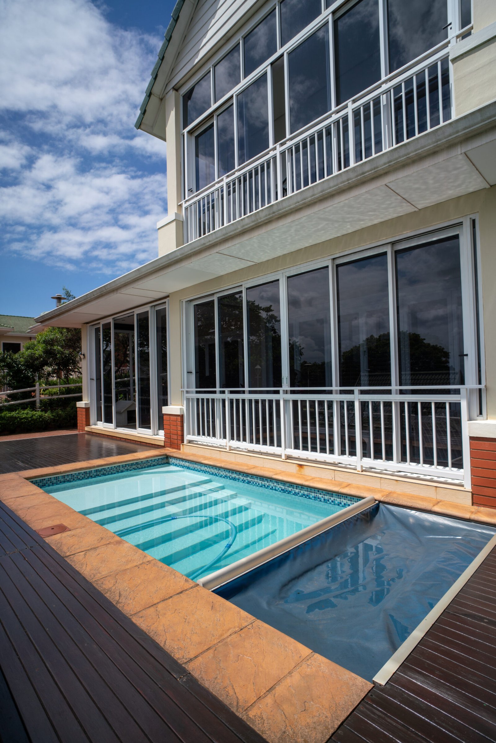 A modern house with large windows overlooks a small rectangular outdoor swimming pool surrounded by a wooden deck and tiled patio.