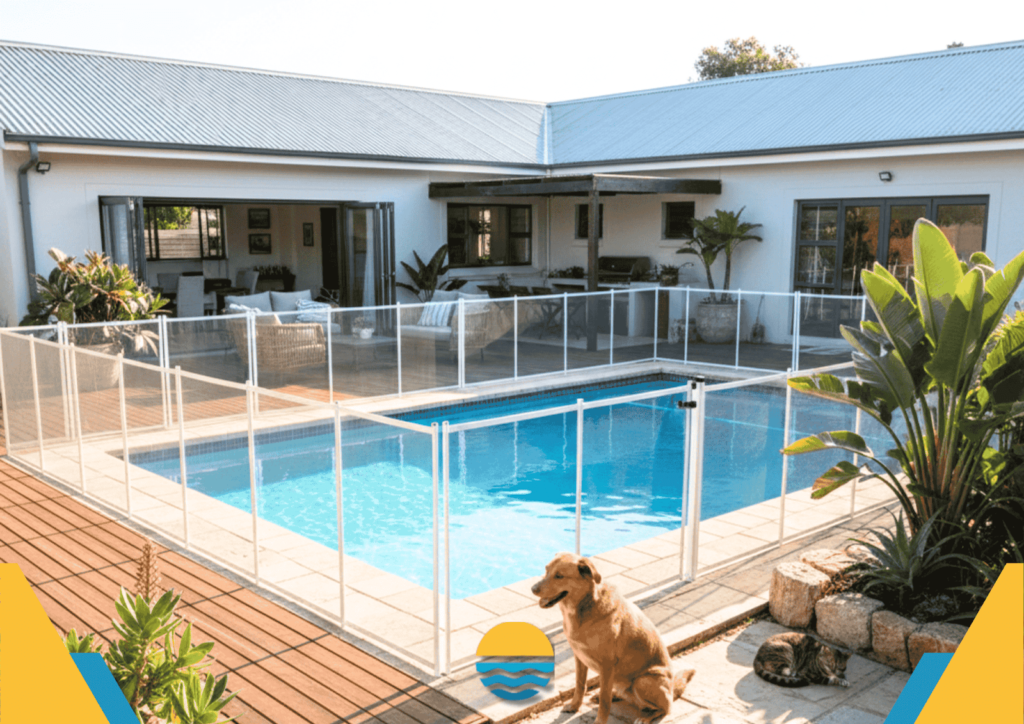 A fenced backyard swimming pool is surrounded by a patio and outdoor furniture. A dog sits on the deck near tropical plants in the foreground.